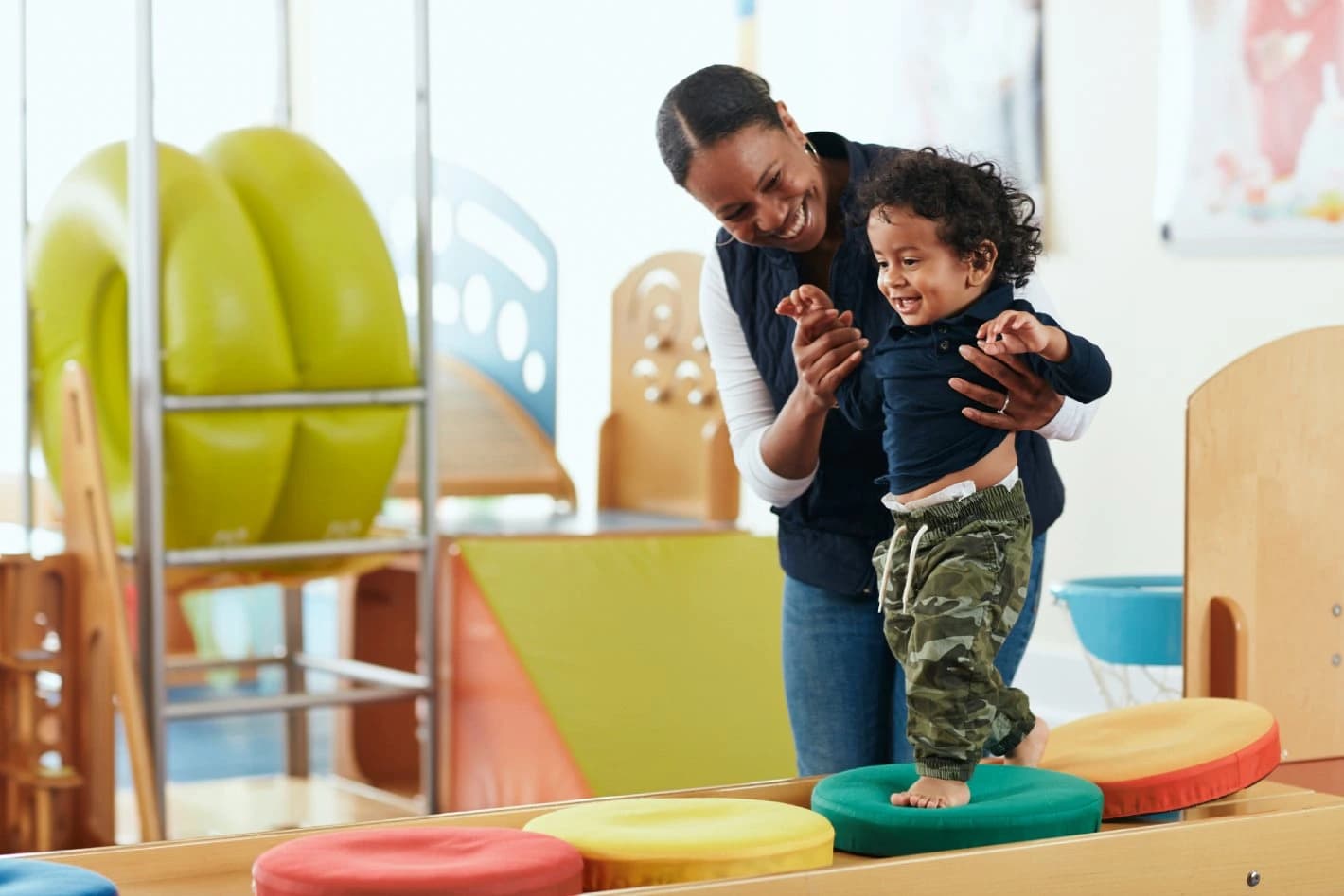 A parent helping a toddler balance on soft stepping stones in a play class