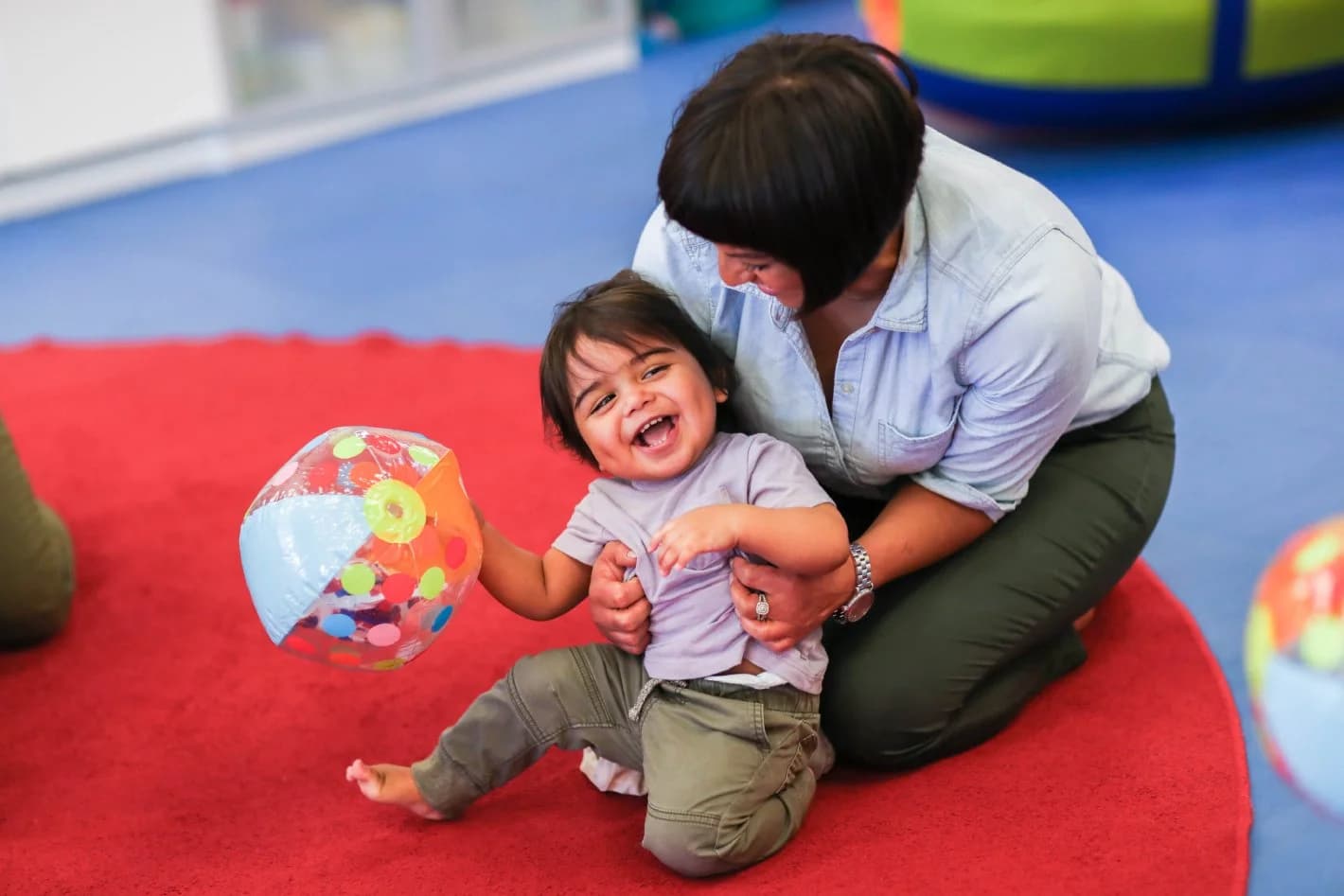 A parent and a laughing toddler playing with a ball on a red play mat